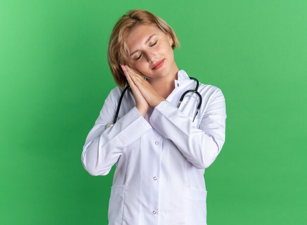 Довольная закрытыми глазами молодая женщина-врач в медицинском халате со стетоскопом, показывающая жест сна, изолированный на зеленой стене
