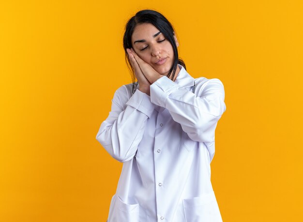 Pleased with closed eyes young female doctor wearing medical robe with stethoscope showing sleep gesture isolated on yellow wall