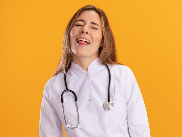 Довольная закрытыми глазами молодая женщина-врач в медицинском халате со стетоскопом показывает язык, изолированный на желтой стене