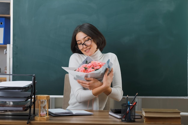 Soddisfatto degli occhi chiusi giovane insegnante seduta alla scrivania con strumenti scolastici che tengono bouquet di fiori in classe