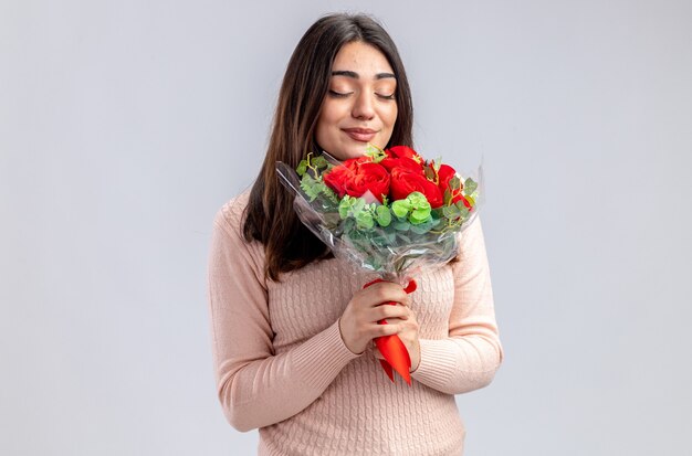 Pleased with closed eyes young girl on valentines day holding bouquet isolated on white background