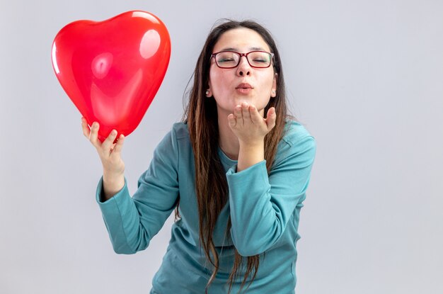 Pleased with closed eyes young girl on valentines day holding heart balloon showing kiss gesture isolated on white background