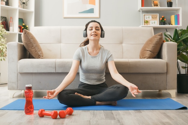 Pleased with closed eyes young girl wearing headphones exercising on yoga mat in front of sofa in living room