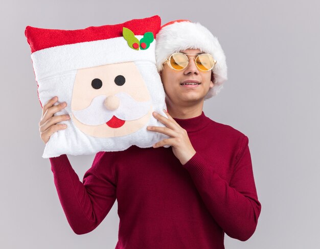 目を閉じて満足している白い背景で隔離のクリスマス枕を保持しているメガネとクリスマス帽子をかぶって若い男