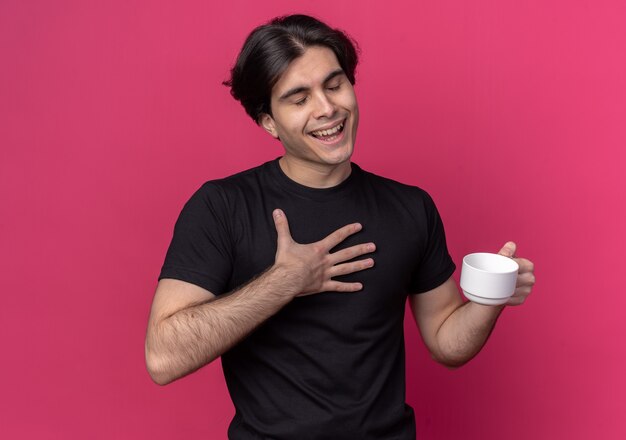 Бесплатное фото Довольный закрытыми глазами молодой красивый парень в черной футболке держит чашку кофе на розовой стене