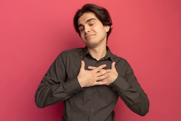 Pleased with closed eyes young handsome guy wearing black t-shirt putting hands on heart isolated on pink wall