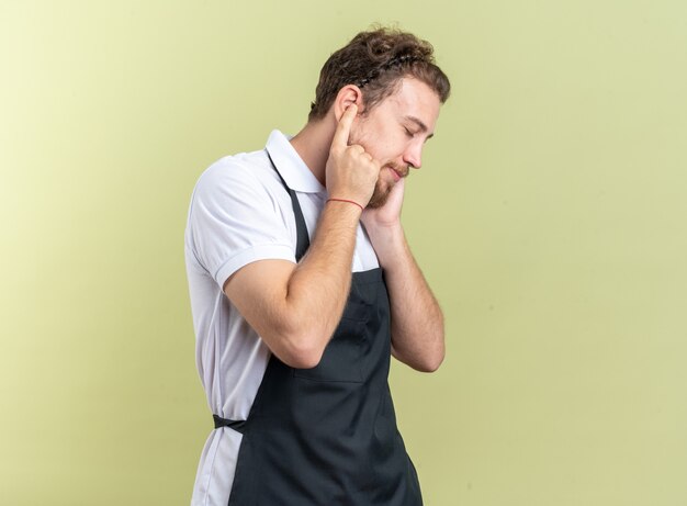 Pleased with closed eyes young male barber wearing uniform covered ears with fingers isolated on olive green wall