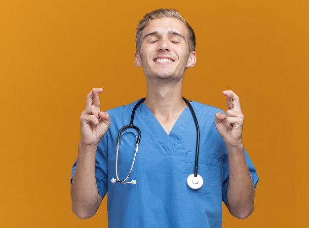 Pleased with closed eyes young male doctor wearing doctor uniform with stethoscope crossing fingers isolated on orange wall