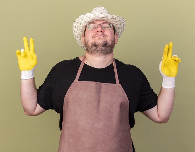 Pleased with closed eyes young male gardener wearing gardening hat and gloves showing peace gesture isolated on olive green wall