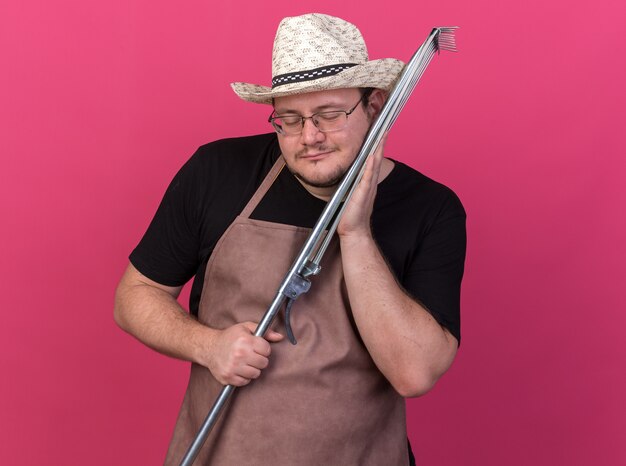 Pleased with closed eyes young male gardener wearing gardening hat holding leaf rake on cheek isolated on pink wall