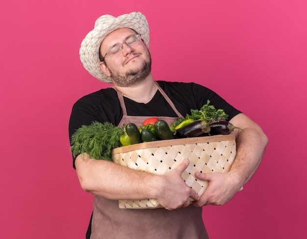 Pleased with closed eyes young male gardener wearing gardening hat holding vegetable basket isolated on pink wall