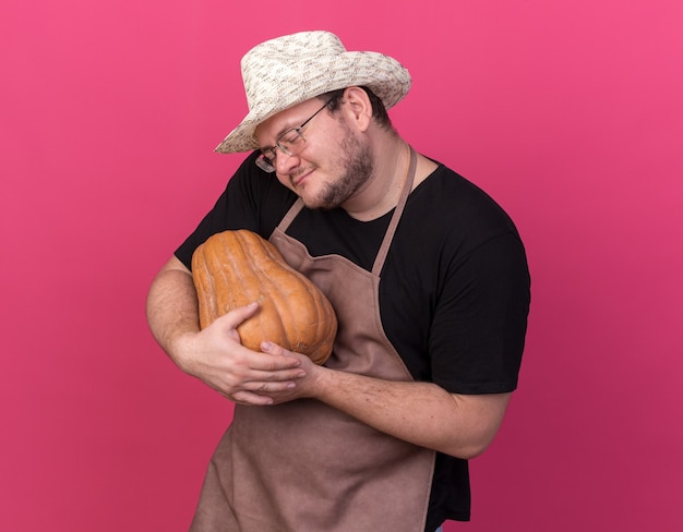 Free photo pleased with closed eyes young male gardener wearing gardening hat hugged pumpkin isolated on pink wall