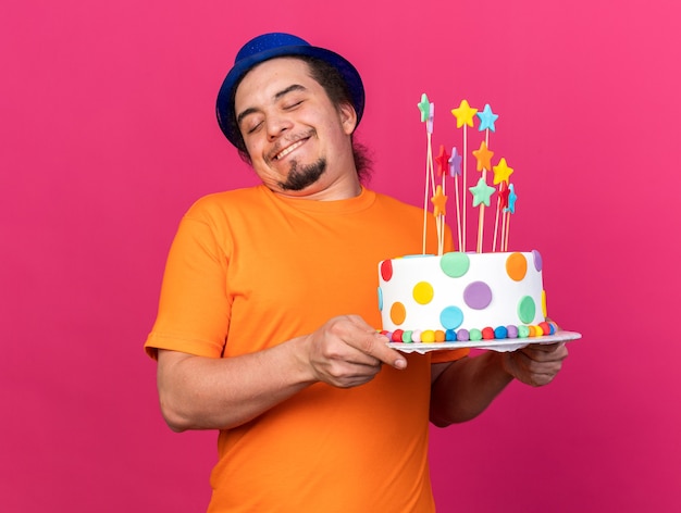 Pleased with closed eyes young man wearing party hat holding cake 