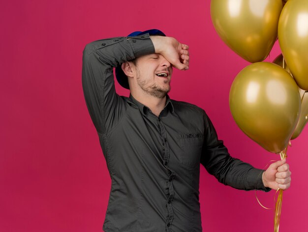 Pleased with closed eyes young party guy wearing blue hat holding balloons putting arm on forehead isolated on pink