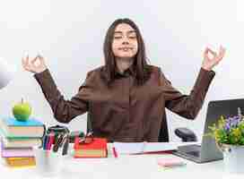 Free photo pleased with closed eyes young school woman sits at table with school tools doing meditation gesture