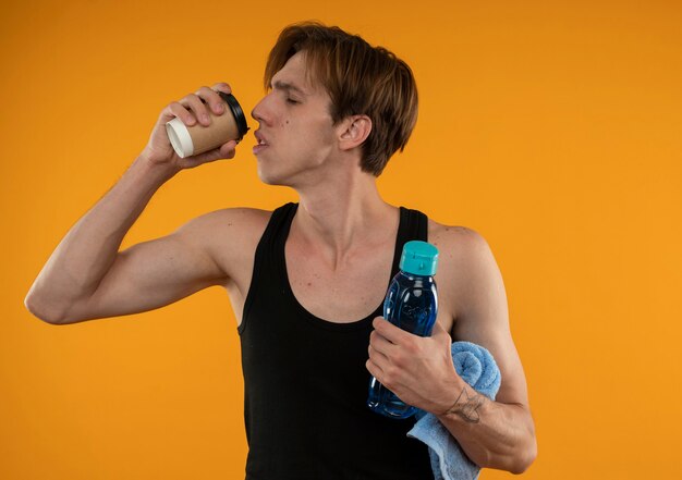 Pleased with closed eyes young sporty guy holding towel with water bottle and drinks coffee isolated on orange wall