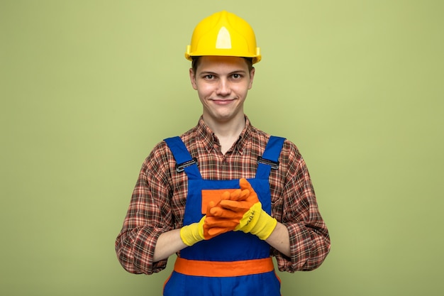 Pleased with hands together young male builder wearing uniform with gloves