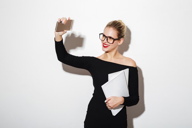 Pleased woman in dress and eyeglasses making selfie on smartphone