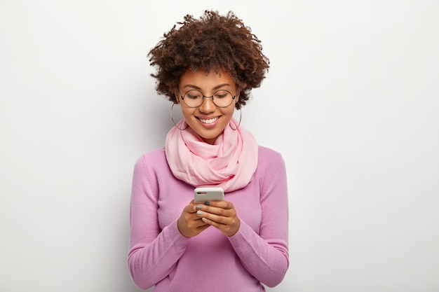Free photo pleased woman focused in smartphone device, has cheerful expression, checks notification or email box, wears glasses and bright clothes