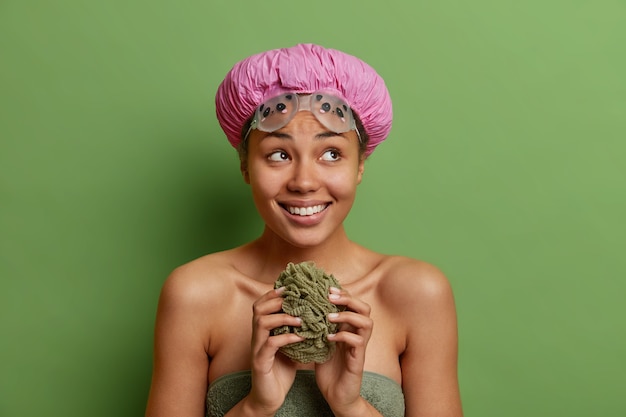 Pleased woman holds sponge going to take shower wears waterproof hat wrapped in bath towel shows bare shoulders isolated over vivid green studio wall