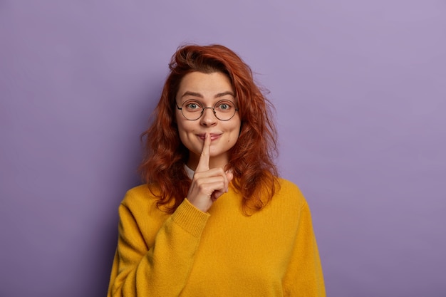 Free photo pleased woman shushing with index finger over lips, tells confidential inforamtion, secret and gossips, wears round glasses