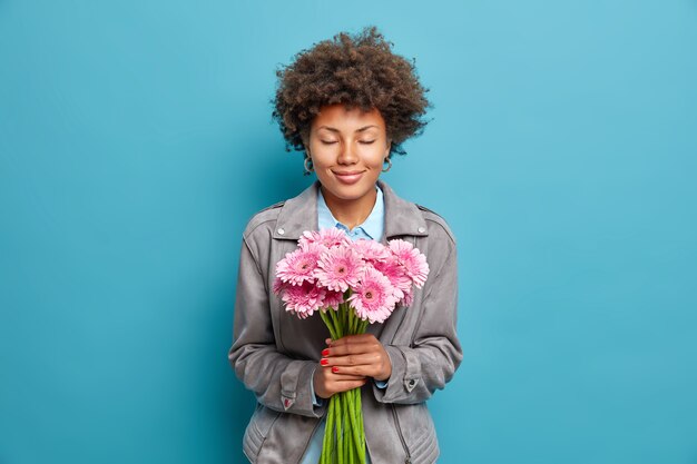 巻き毛の満足している女性は目を閉じて保持します美しいピンクのガーベラの花は青い壁に隔離された灰色のジャケットに身を包んだお祝いの日を楽しんでいます