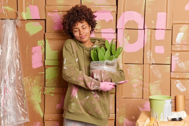 Довольная молодая афроамериканка наслаждается ремонтом дома, намазанная красками, держит кисть, кактус в горшке переезжает в новую квартиру