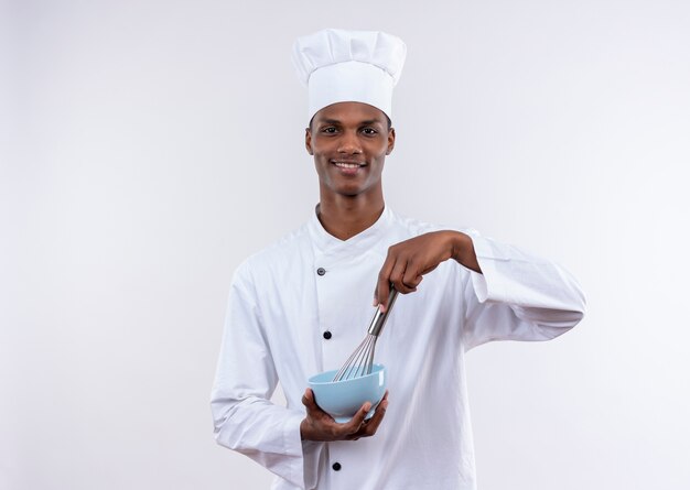 Pleased young afro-american cook in chef uniform holds bowl and whisk on isolated white background with copy space