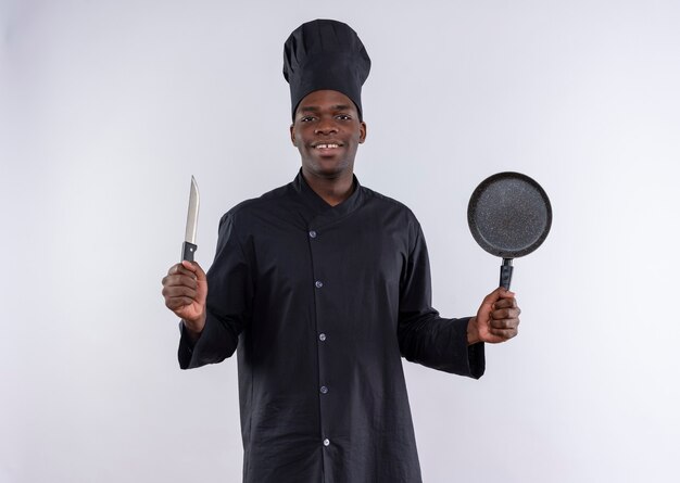 Довольный молодой афро-американский повар в униформе шеф-повара держит нож и сковороду на белом с копией пространства