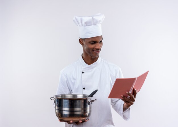 Pleased young afro-american cook in chef uniform holds saucepan and looks at notebook on isolated white background with copy space