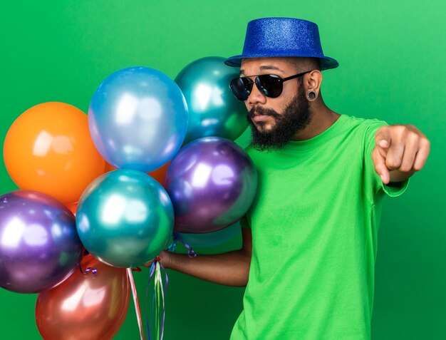 Довольный молодой афро-американский парень в шляпе и очках держит воздушные шары спереди, изолированные на зеленой стене