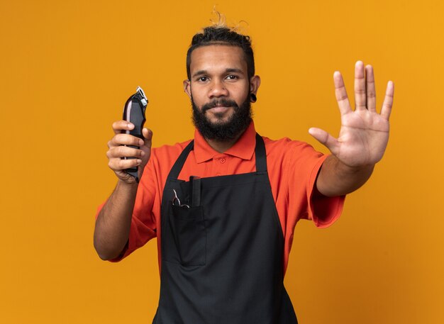 Pleased young afro-american male barber wearing uniform holding hair clippers doing stop gesture 