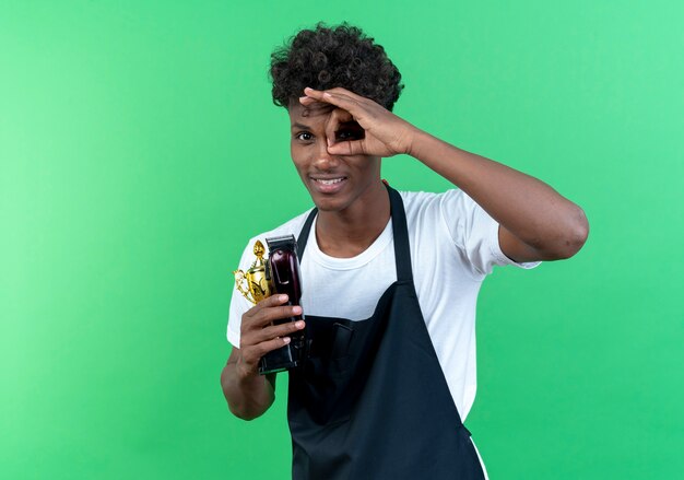 Pleased young afro-american male barber wearing uniform holding hair clippers and showing look gesture isolated on green background