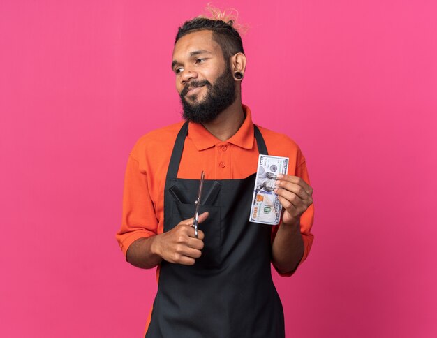 Pleased young afro-american male barber wearing uniform holding scissors and dollar looking at side 