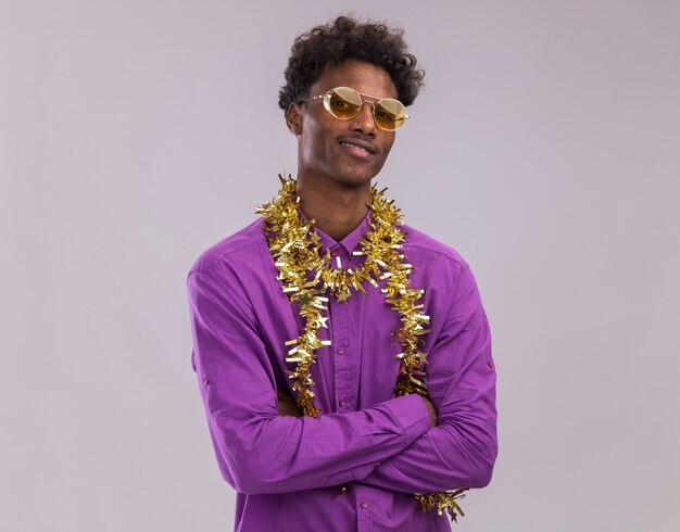 Pleased young afro-american man wearing glasses with tinsel garland around neck standing with closed posture looking at camera isolated on white background