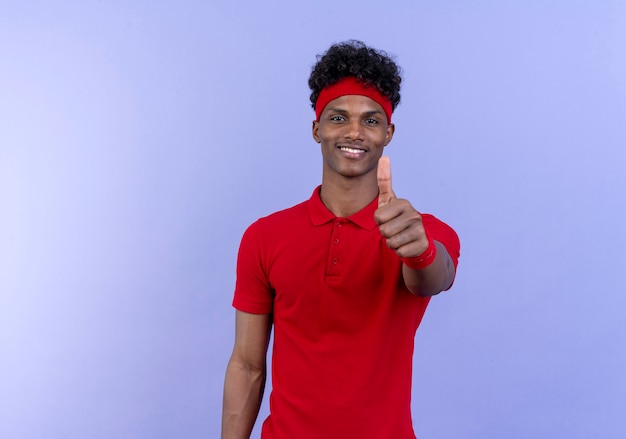 Довольный молодой афро-американский спортивный мужчина, носящий повязку на голову и браслет, подняв большой палец вверх, изолирован на синем фоне