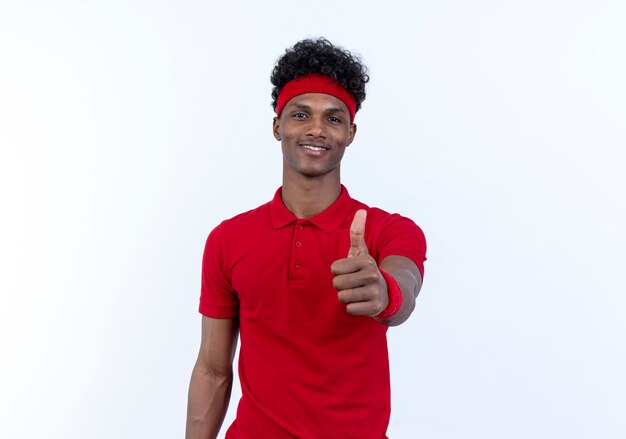 Довольный молодой афро-американский спортивный мужчина с повязкой на голову и браслет, подняв большой палец вверх, изолирован на белом фоне