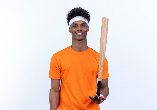 Pleased young afro-american sporty man wearing headband and wristband holding bat on hand isolated on white background