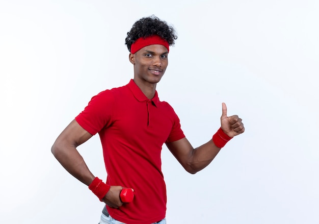 Pleased young afro-american sporty man wearing headband and wristband holding dumbbell around hip his thumb up isolated on white background