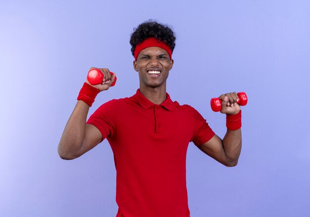 Довольный молодой афро-американский спортивный мужчина с головной повязкой и браслетом, держащий гантели на синей стене