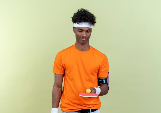 Pleased young afro-american sporty man wearing headband and wristband holding and looking at ping pong racket with ball isolated on green background