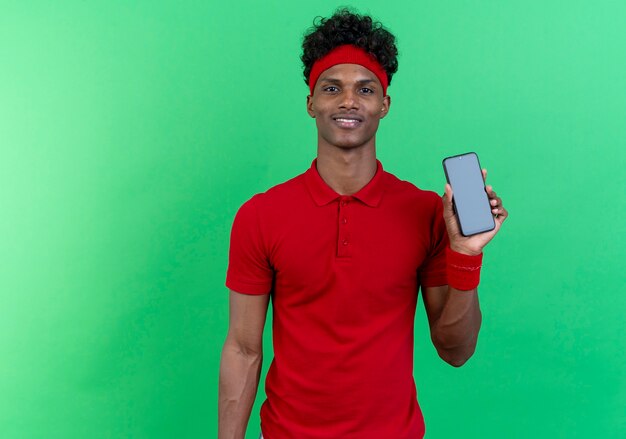 Довольный молодой афро-американский спортивный мужчина с повязкой на голову и браслетом, держащим телефон, изолированным на зеленой стене