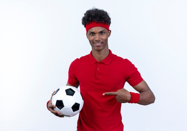 Pleased young afro-american sporty man wearing headband and wristband holding and points at ball isolated on white background
