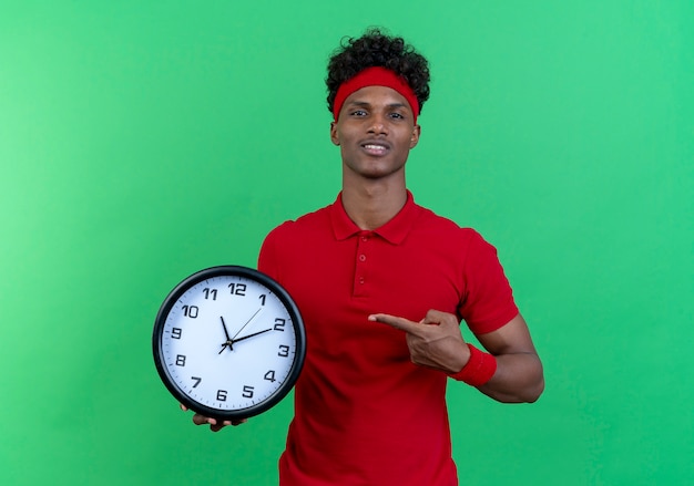 Pleased young afro-american sporty man wearing headband and wristband holding and points at wall clock isolated on green background