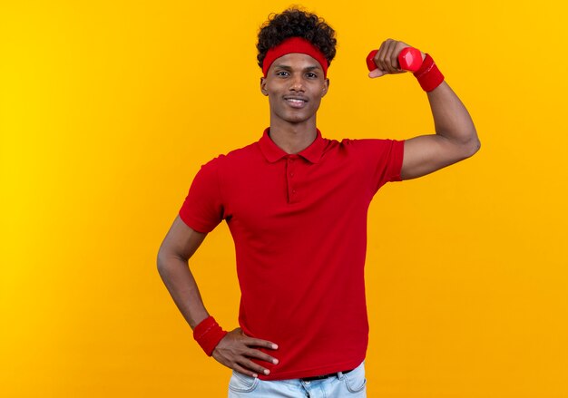 Pleased young afro-american sporty man wearing headband and wristband raising dumbbell and putting hand on hip isolated on yellow background