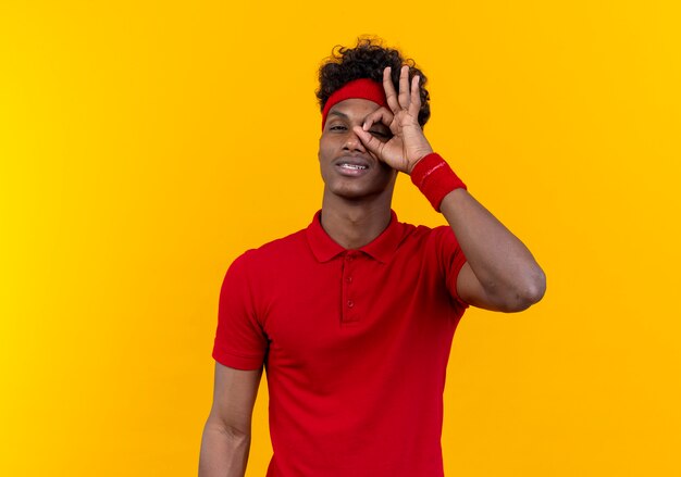 Pleased young afro-american sporty man wearing headband and wristband showing look gesture isolated on yellow background