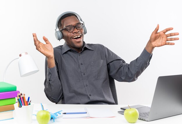 Pleased young afro-american student in optical glasses and on headphones sitting at desk with school tools keeping hands open isolated on white wall