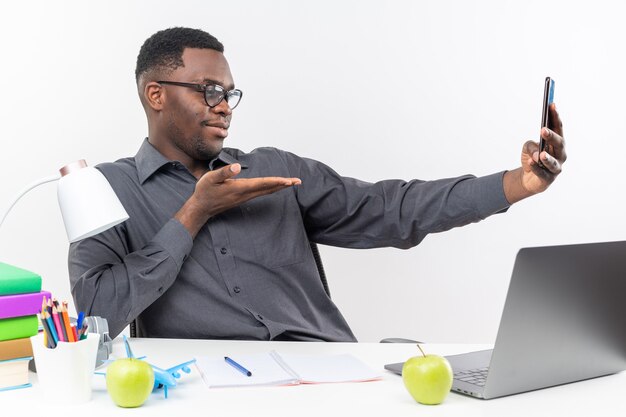 Pleased young afro-american student in optical glasses sitting at desk with school tools looking