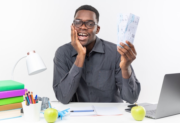 Pleased young afro-american student in optical glasses sitting at desk with school tools putting hand on his face and holding air tickets isolated on white wall