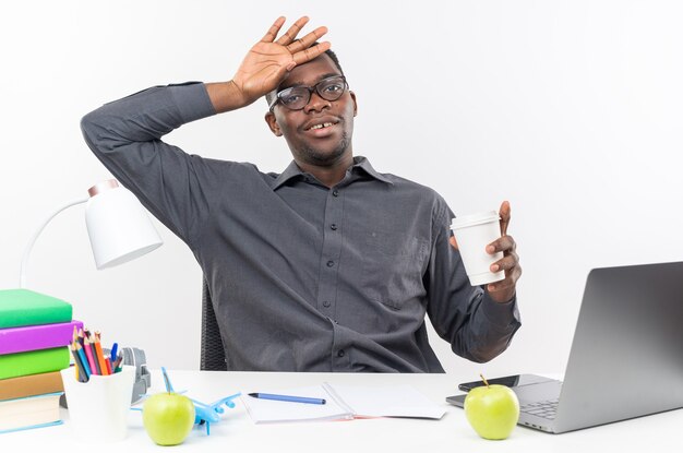 Pleased young afro-american student in optical glasses sitting at desk with school tools putting hand on his head and holding paper cup isolated on white wall